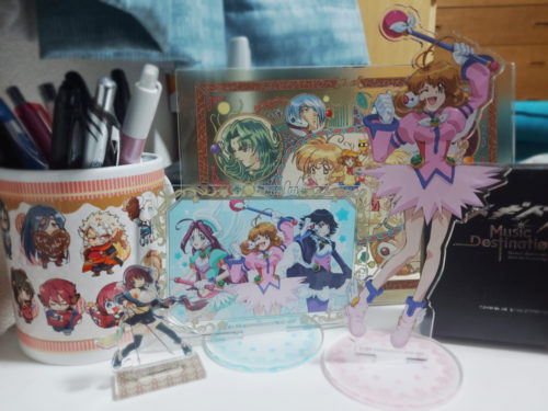 グッズがごちゃごちゃ並んでいる写真。コレユイのアクスタ、パネル、うたわれのマグカップ、メギドの時計など